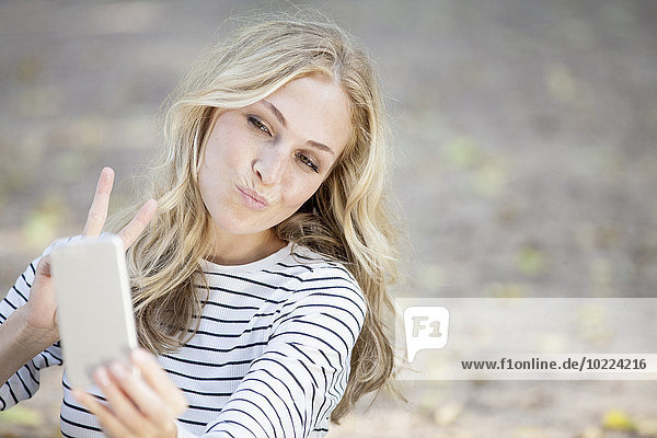 Porträt einer lächelnden blonden Frau  die einen Selfie mit ihrem Smartphone nimmt.