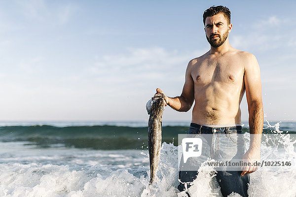 Portrait of young man standing in the water of the sea holding caught fish
