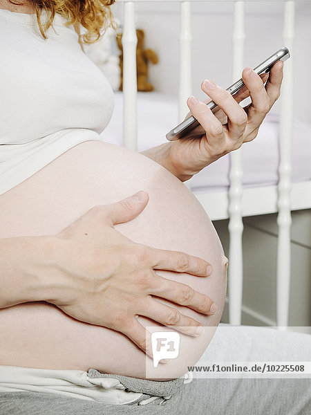 Schwangere Frau vor dem Kinderbett mit einem Smartphone