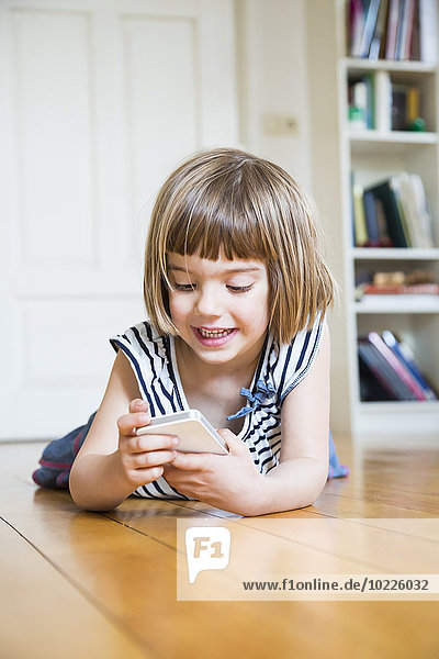 Portrait of smiling little girl lying on wooden floor with smartphone