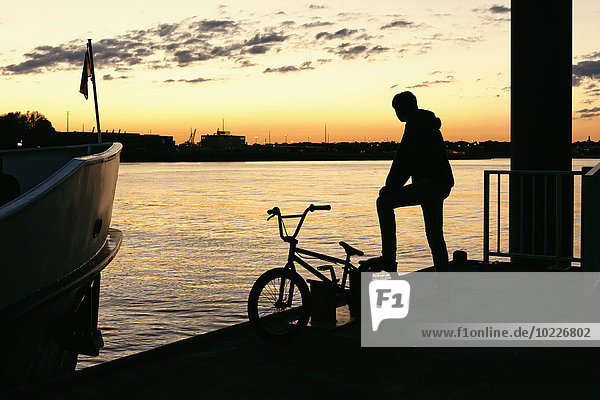 Deutschland,  Hamburg,  Silhouette eines jungen Mannes mit Fahrrad am Steg bei Abendlicht
