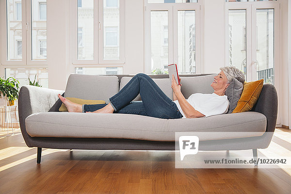 Reife Frau entspannt mit digitalem Tablett auf der Couch im Wohnzimmer