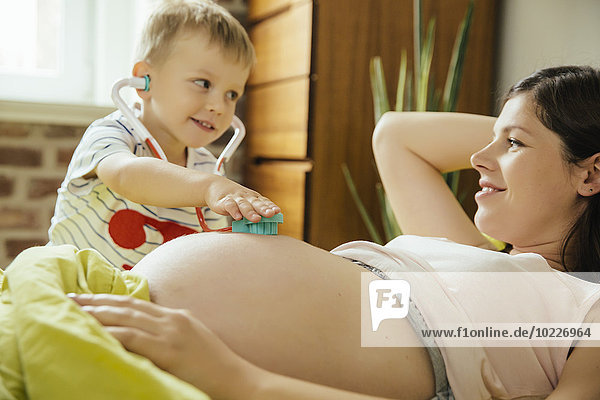 Kleiner Junge benutzt ein Spielzeug-Stethoskop  um den Bauch seiner schwangeren Mutter zu untersuchen.