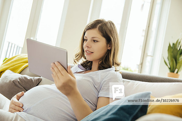 Pregnant woman with digital tablet relaxing on couch at home