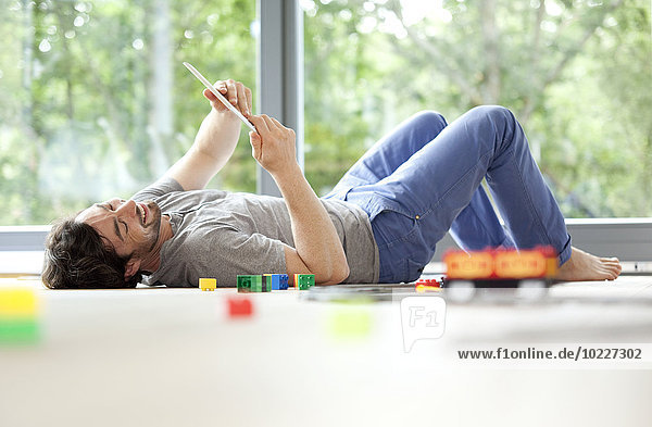 Lächelnder Mann auf dem Boden liegend mit digitalem Tablett neben der Spielzeugeisenbahn