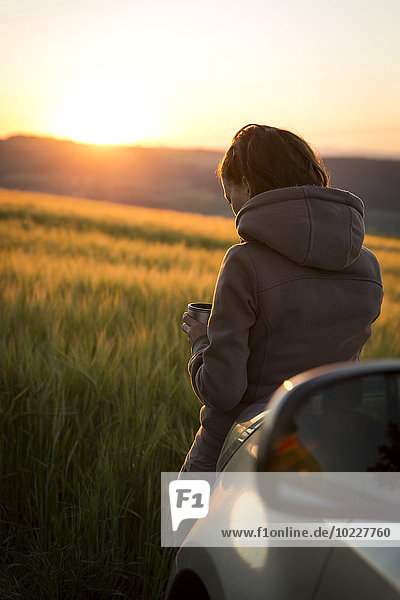 Deutschland  Frau am Auto vor einem Feld bei Sonnenaufgang