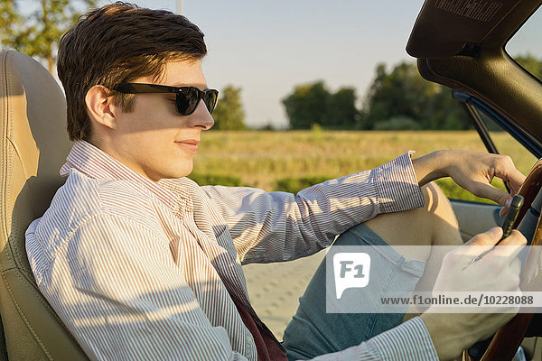 Teenager mit Sonnenbrille schaut auf sein Smartphone in einem Cabriolet