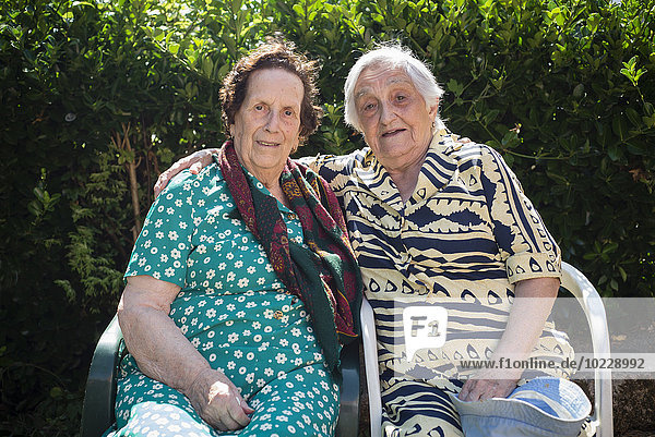 Porträt von zwei älteren Frauen  die nebeneinander auf Stühlen im Garten sitzen.