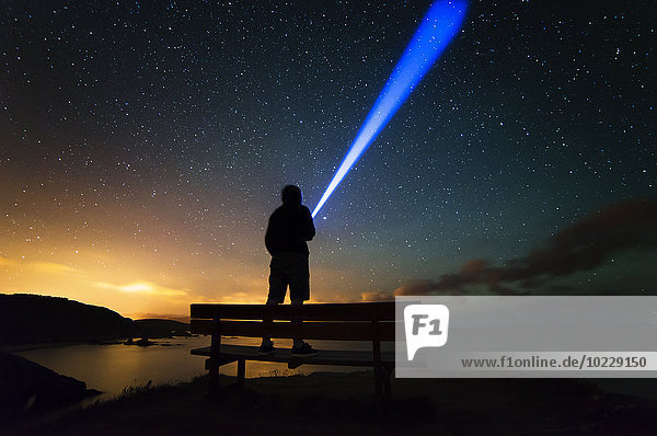 Spanien  Ortigueira  Loiba  Silhouette eines Mannes auf der Bank unter dem Sternenhimmel mit blauem Strahl