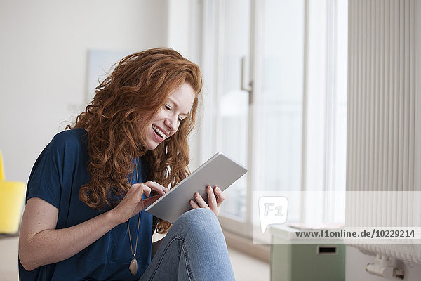 Fröhliche junge Frau sitzt in ihrem Wohnzimmer mit digitalem Tablett