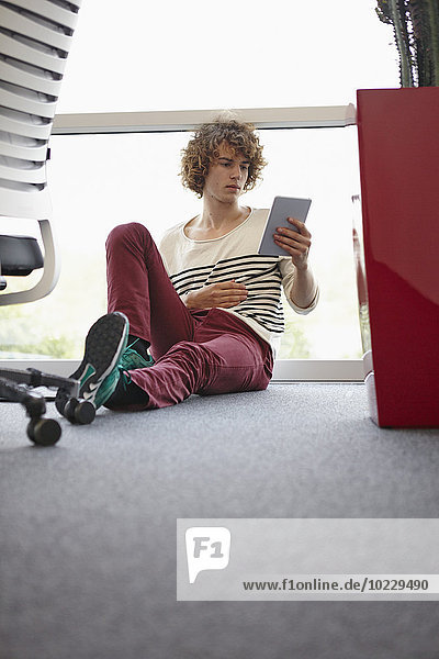 Junger Mann im Büro sitzend auf dem Boden mit digitalem Tablett
