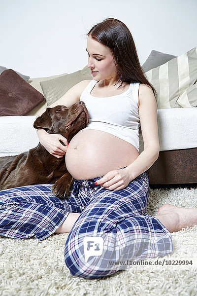 Schwangere Frau sitzt mit ihrem Labrador Retriever auf dem Boden ihres Wohnzimmers.