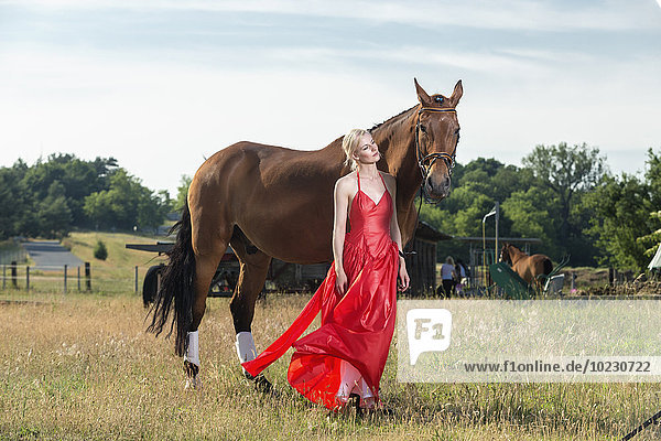 Porträt einer jungen Frau in rotem Abendkleid neben einem Pferd auf einer Wiese stehend