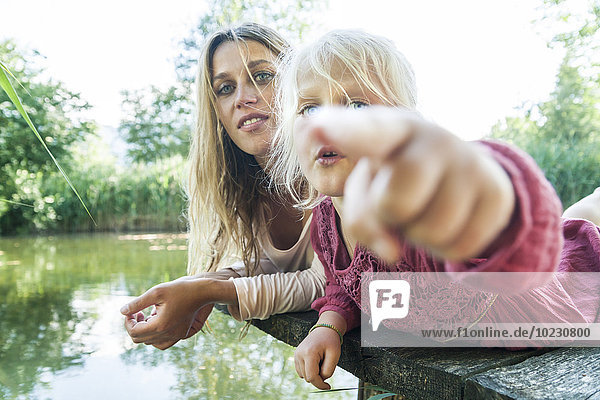 Girl lying with mother on jetty at a lake pointing finger