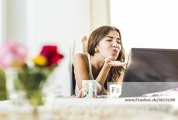 Relaxed young woman lying in bed using laptop blowing a kiss