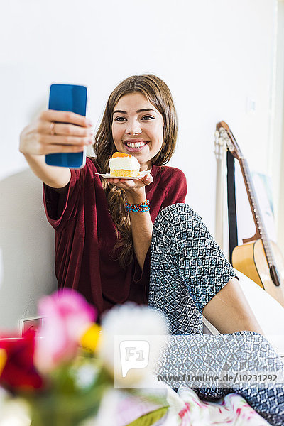 Entspannte junge Frau zu Hause im Bett  die Selfie mit einem Stück Kuchen nimmt.