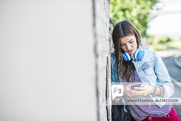 Brünette junge Frau mit Kopfhörer und Handy gegen die Wand gelehnt