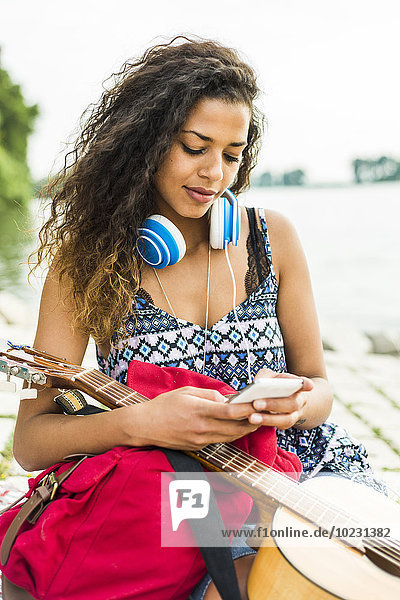 Junge Frau mit Kopfhörer  Gitarre  Handy und Rucksack am Flussufer