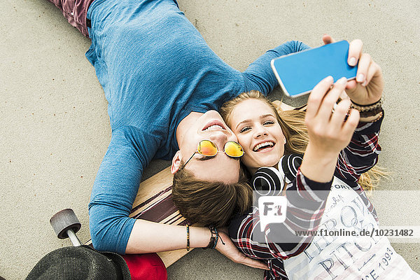 Glückliches junges Paar auf dem Boden liegend mit Skateboard und Blick auf Handy