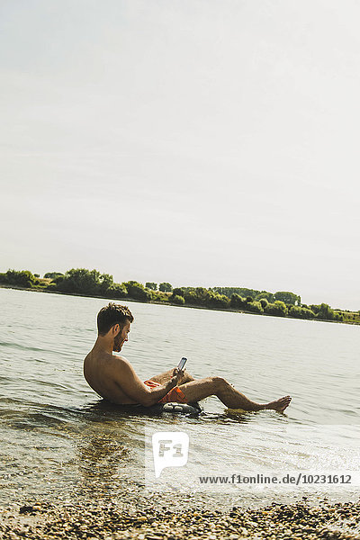 Junger Mann sitzt in einer Röhre im Fluss und benutzt ein Smartphone.