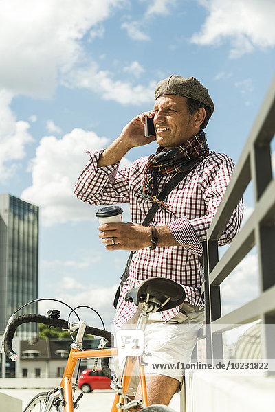 Erwachsener Mann mit Fahrrad an die Wand gelehnt  mit Kaffeetasse und Smartphone