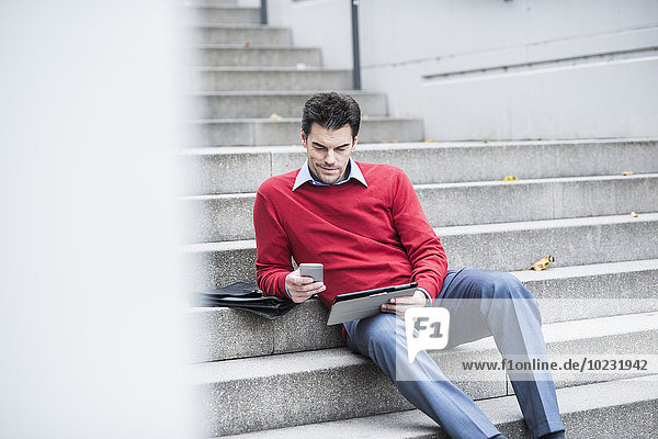 Geschäftsmann mit digitalem Tablett und Smartphone auf der Treppe sitzend