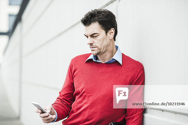 Porträt eines Geschäftsmannes  der sich an eine Betonwand lehnt und sein Smartphone betrachtet