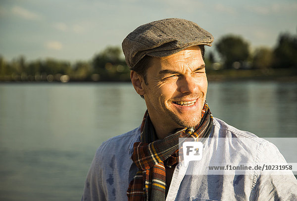 Porträt eines lächelnden Mannes mit Mütze vor einem Fluss in der Abenddämmerung.