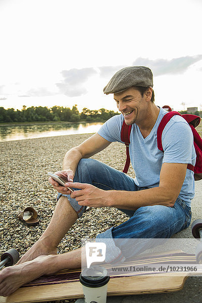Lächelnder Mann am steinigen Strand mit Skateboard und Smartphone am Abend