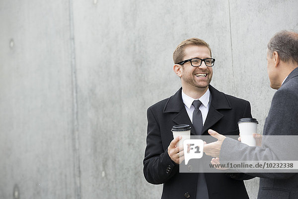 Two smiling businessmen with coffee to go at concrete wall