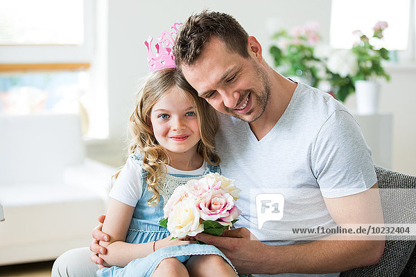 Mädchen mit rosa Krone auf Vaters Schoß sitzend mit Blumenstrauß
