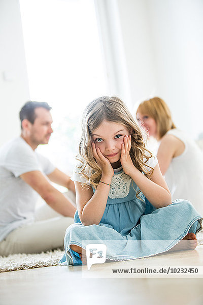 Little girl sitting cross-legged on floor  parents talking in background