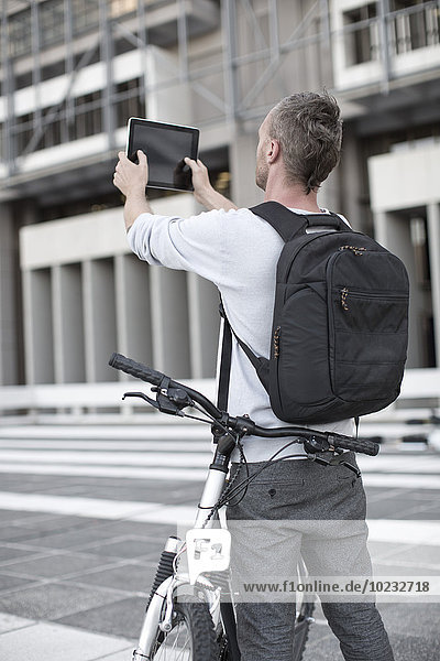 Man with rucksack and bike taking picture with digital tablet
