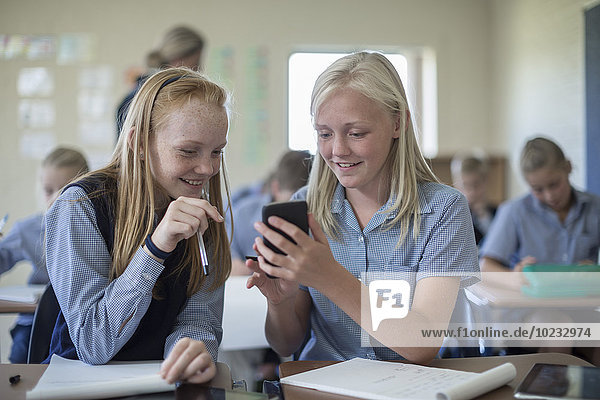 Two smiling schoolgirls in classroom looking at cell phone
