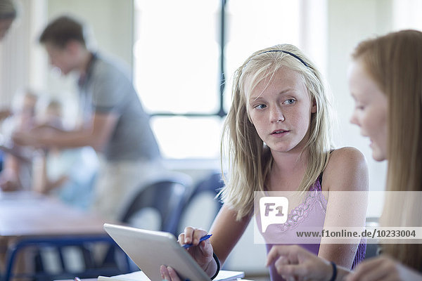 Two schoolgirls in classroom with digital tablet talking