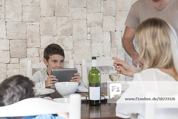 Familie beim Mittagessen am Esstisch mit einem Jungen  der ein digitales Tablett hält.