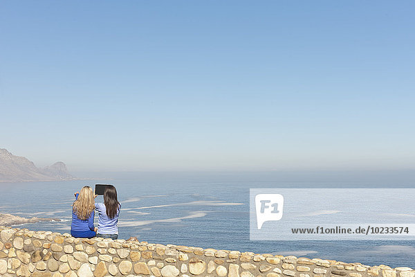 South Africa  two women sittin on a wall at the coast taking a picture with digital tablet