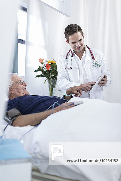 Doctor showing senior man lying in hospital bed something on digital tablet
