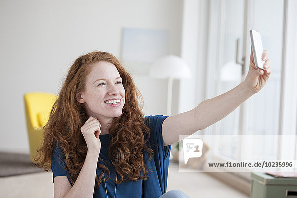 Junge Frau sitzt in ihrem Wohnzimmer und nimmt einen Selfie mit dem Smartphone.