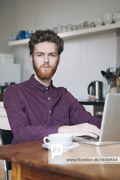 Portrait of confident young man using laptop at table in kitchen