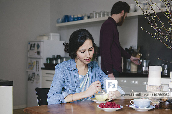 Young woman stirring coffee at kitchen table with man in background