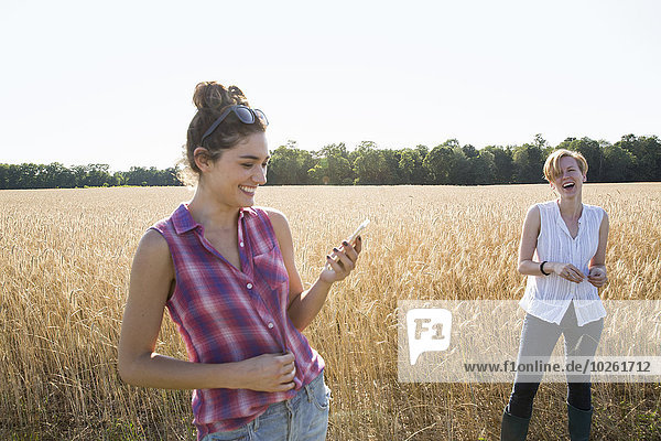 Zwei junge Frauen stehen in einem Maisfeld  eine hält ihr Handy in der Hand.