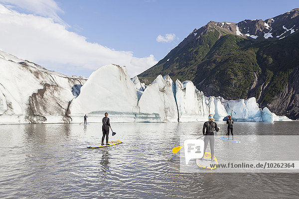 Group of four people stand up paddleboarding in front of Grewingk Glacier  Kachemak Bay State Park  Southcentral Alaska