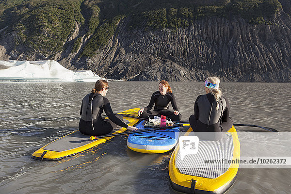 Three women sitting on top of their inflatable stand up paddle boards eating a snack in front of the Grewingk glacier in Kachemak Bay State Park  Southcentral Alaska.