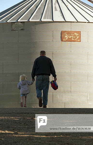 A farmer and his young daughter walk hand-in-hand through his farm yard with a grain bin in the background  near Sioux City; Iowa  United States of America