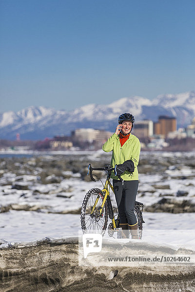 Young woman with her bicycle talks on a cell phone while standing on frozen ice chunks along the Tony Knowles Coastal Trail  Anchorage  Southcentral Alaska