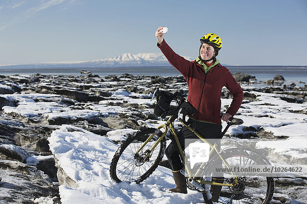Young woman with her bicycle takes a selfie with a smart phone while standing on frozen ice chunks along the Tony Knowles Coastal Trail  Anchorage  Southcentral Alaska