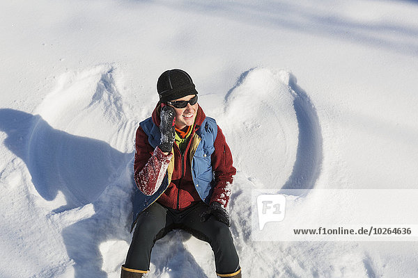A smiling young woman wearing a hat and gloves talks on her cell phone after making a snow angel in a snowbank  Winter  Southcentral Alaska  USA