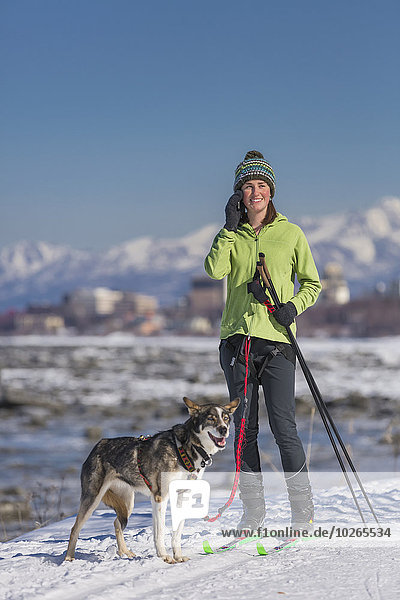 A young woman stops and talks on a cell phone while she skijors with her Alaska Husky sled dog down the Tony Knowles Coastal Trail  Anchorage  Southcentral Alaska  USA.