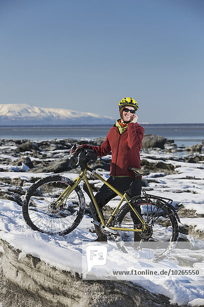 A young woman stands with her bicycle on ice while talking on a cell phone next to the Tony Knowles Coastal Trail  Anchorage  Southcentral Alaska  USA.
